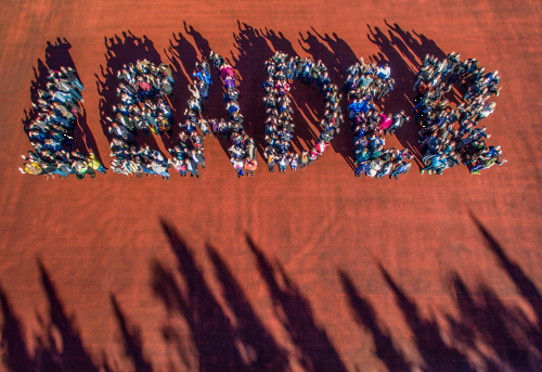 Das Bild zeigt eine Menschengruppe. Diese hat sich in kleinere Gruppen aufgeteilt. In den kleinen Gruppen haben sich die Menschen so angeordnet, dass jeweils eine der Gruppen einen Buchstaben bildet. Alle Gruppen zusammen ergeben das Wort LEADER. Das Bilde ist eine Draufsicht auf das Wort, sodass es lesbar wird. Zudem stehen die Menschen auf einem roten Untergrund, wie man ihn von Sportplätzen kennt und im Unteren Bildrand ragen Schatten von vermutlich Bäumen in das Bild.